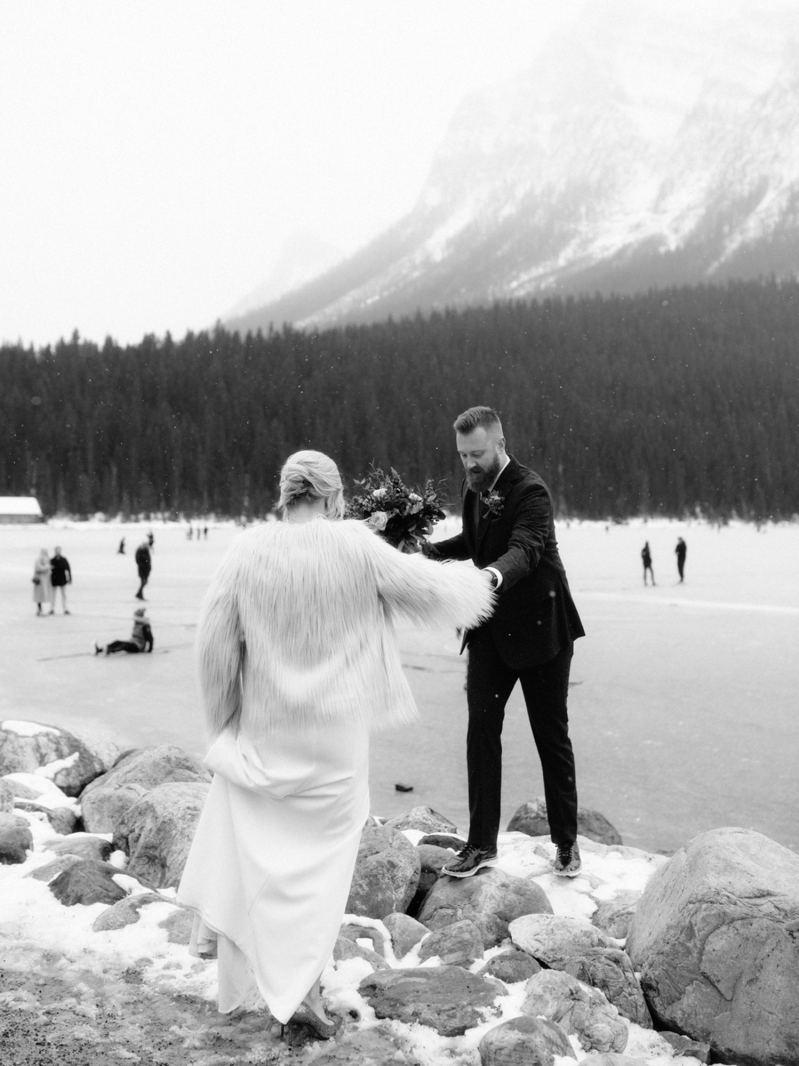 Groom helping his new bride up on rocks alongside the frozen glacial Lake Louise with hockey players behind them