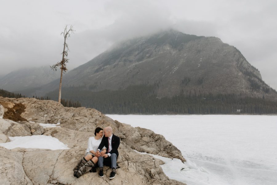 Couple snuggling on Lake Minnewanka rocks, she is wearing traditional mukluks and a Ted Baker dress