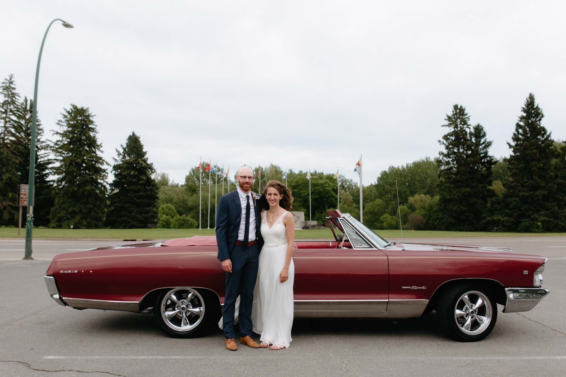 Couple posing beside a maroon 1960s getaway car at the Confederation Park parking lot
