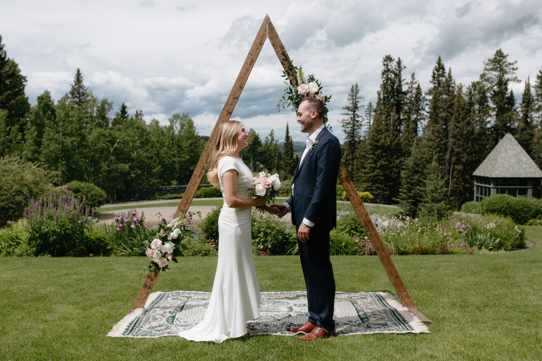 Small wedding ceremony setup with couple holding hands while stnading on elaborate rug and under a triangle arch with florals