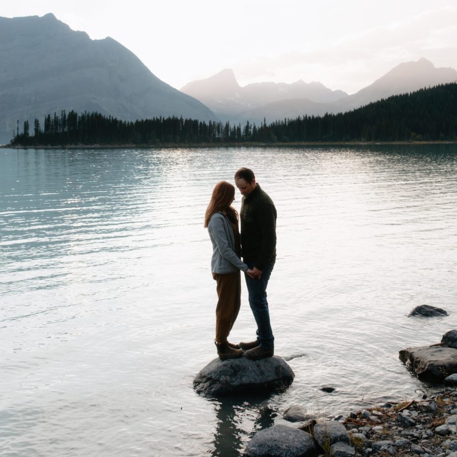 Anniversary photography session at Upper Kananaskis Lakes with couple embracing on a rock in the lack with hazy mountain skies behind them