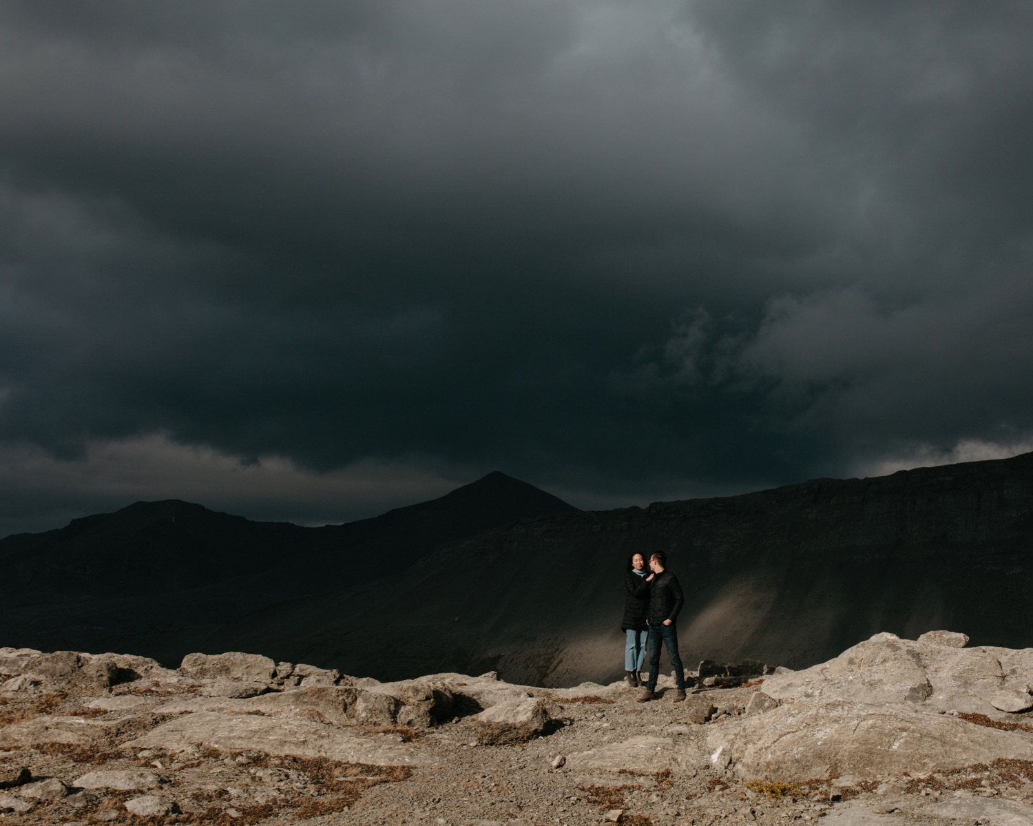 Moody skies overhead casting light spots on the mountain behind a couple standing on a rock ridge in Jasper