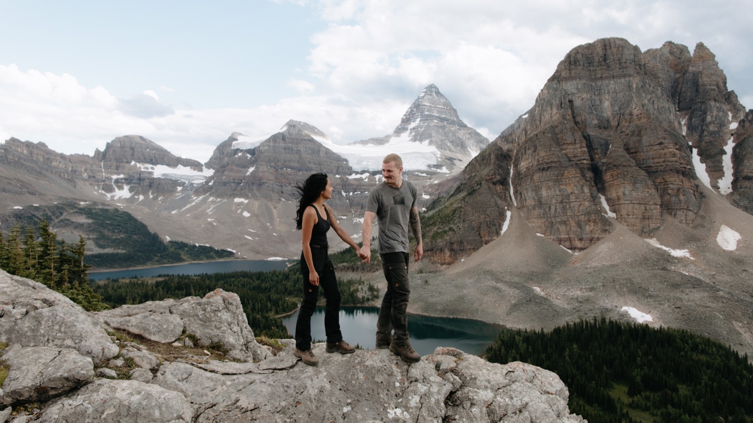 Mountainous and glacier fed backdrop as couple hold hands hiking across a rock ledge above Sunburst Lake
