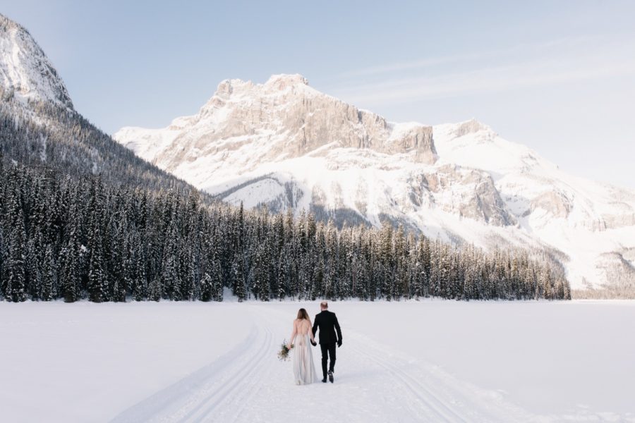 Newly married couple wearing backless wedding dress walking across a frozen Emerald Lake for their February winter wedding