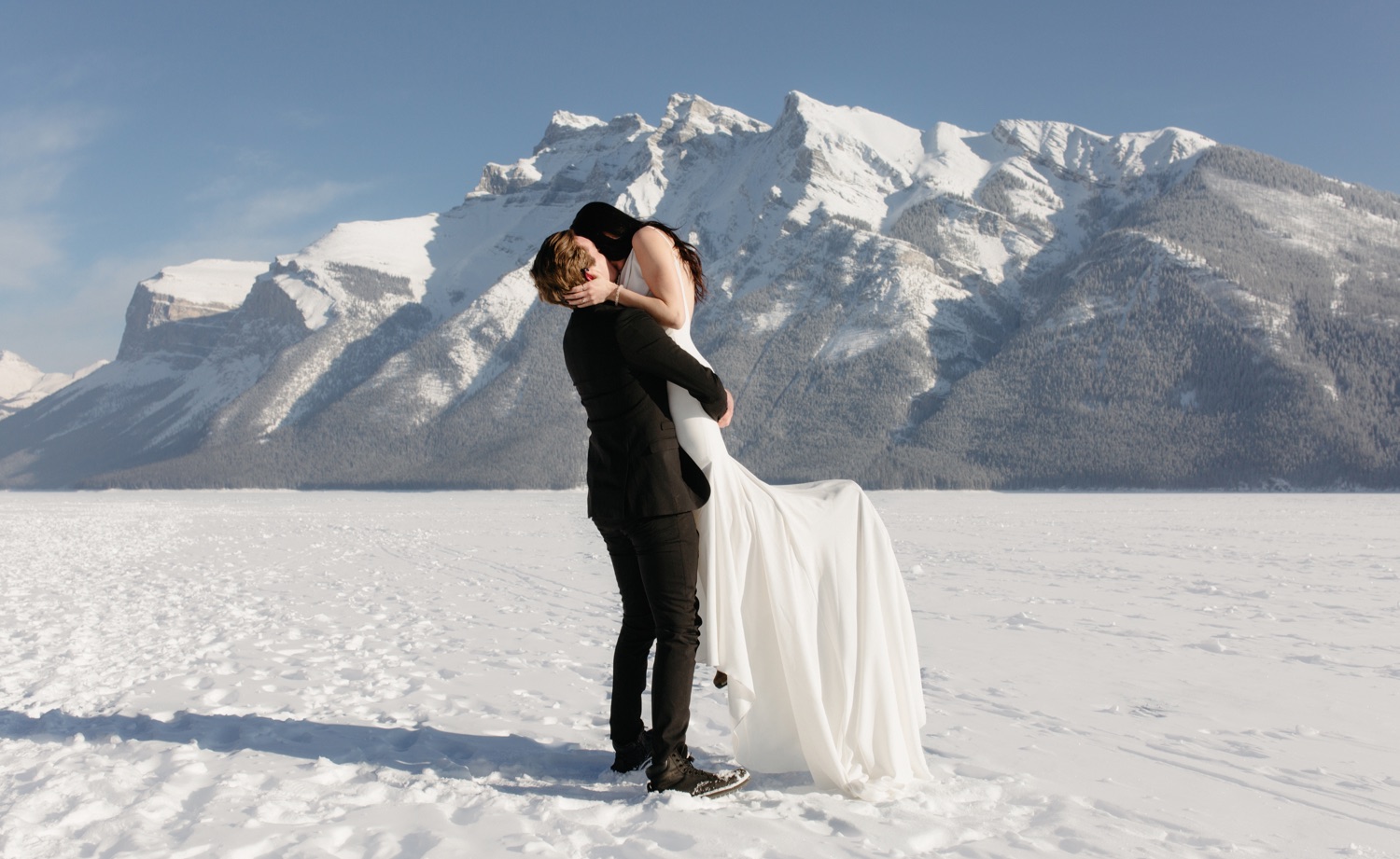 Newly married couple embracing on frozen Lake Minnewanka with soft warm white hues and soft blue sky