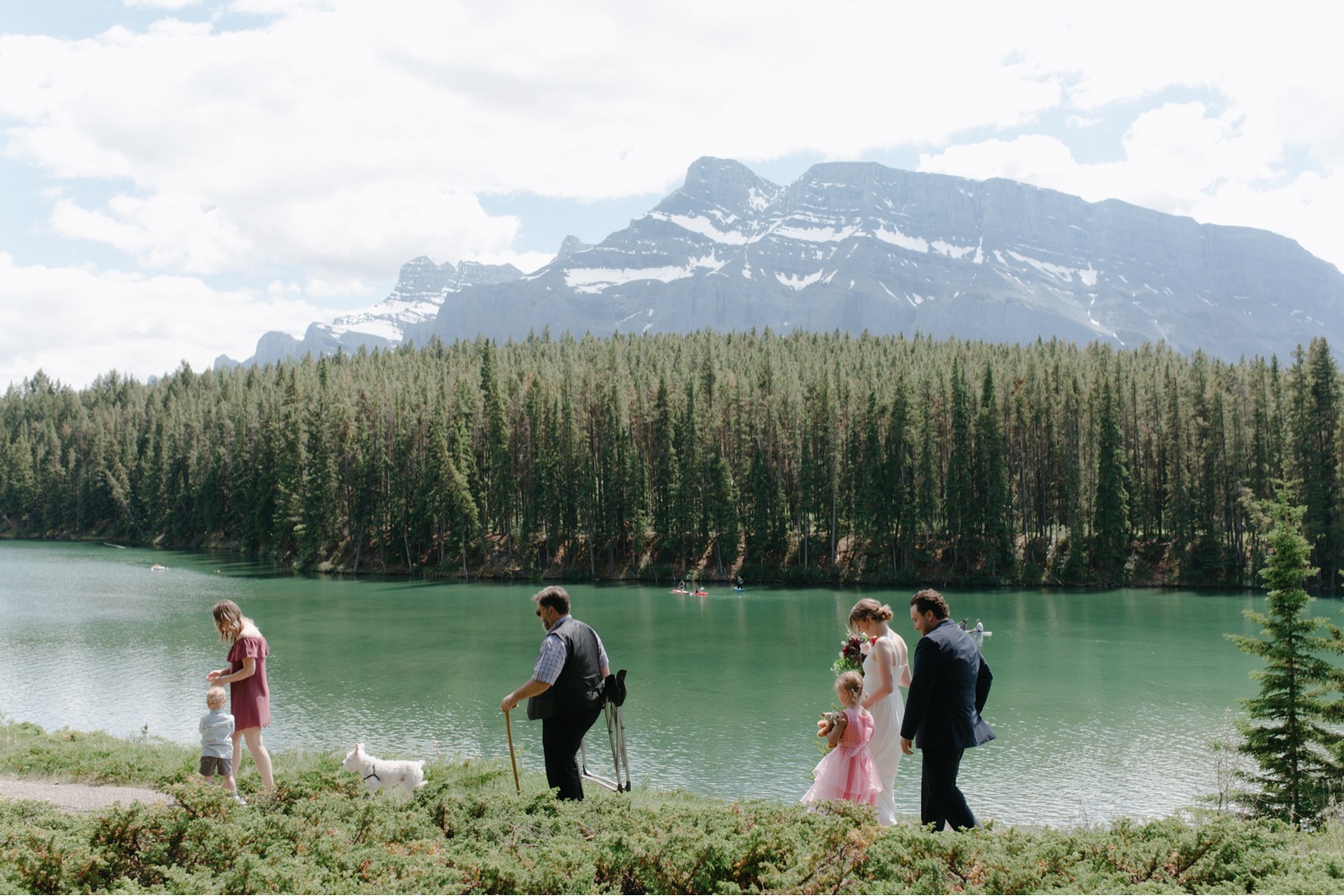 Wedding guests carrying chairs along the Johnson Lake shore with green water and Mount Rundle backdrop