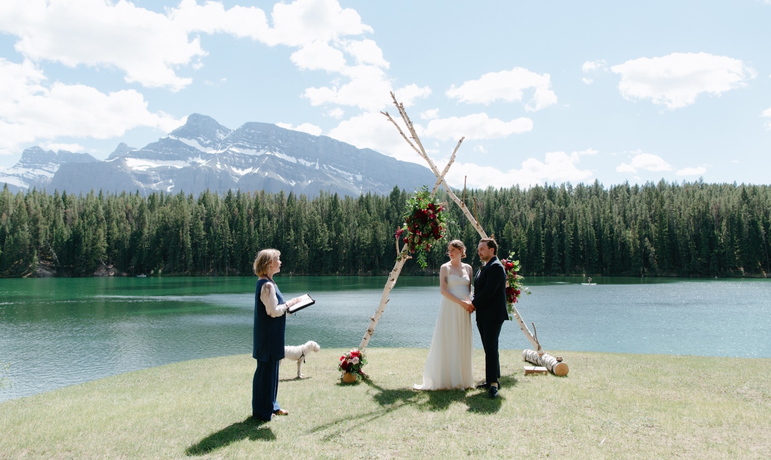 Intimate wedding ceremony at Johnson Lake, with couple standing under birch altar with mahogany florals, as Barbara Parker officiates