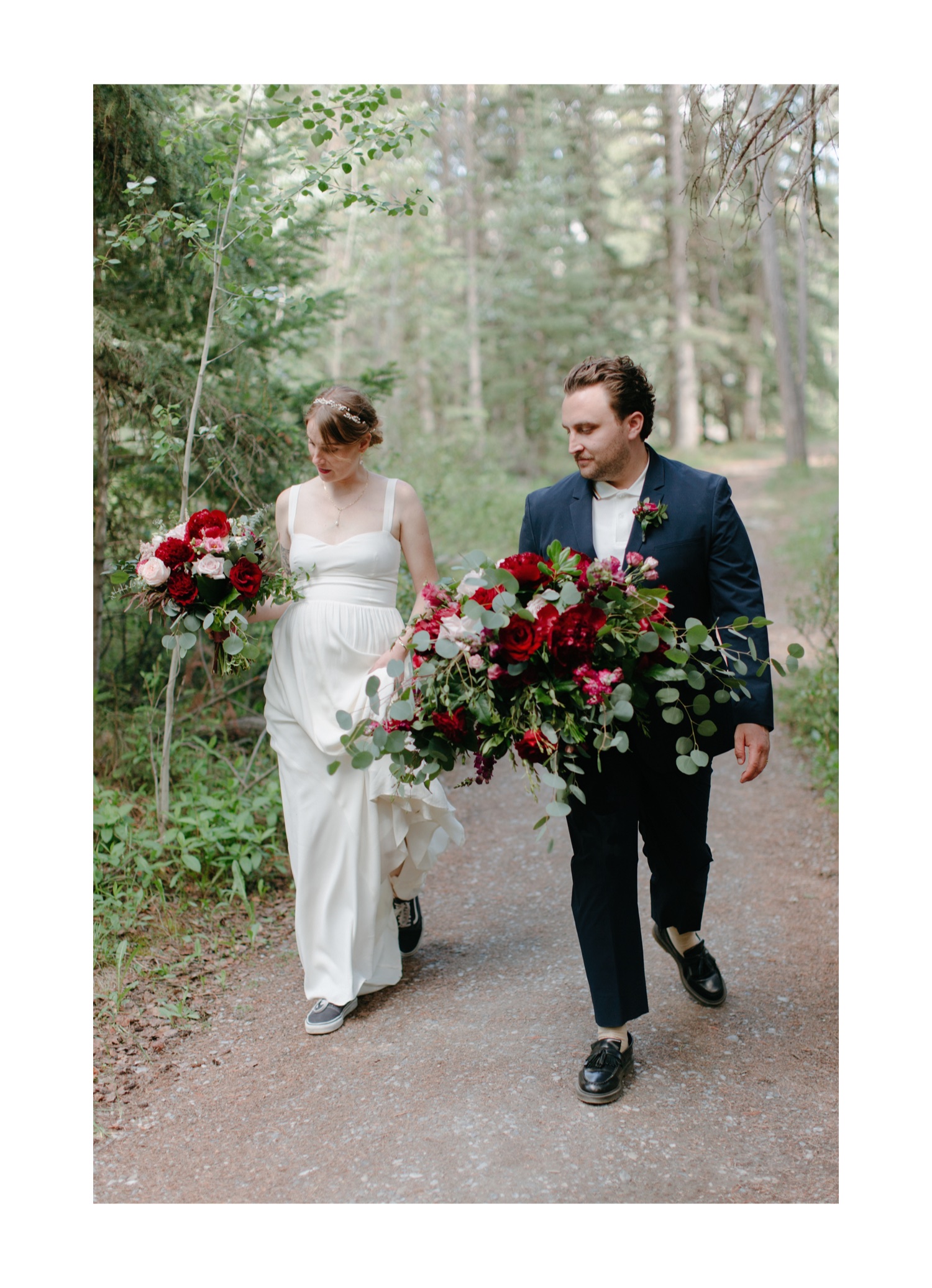 Married couple wearing Reformation Dress and Hugo Boss suit carrying red, pink, white, and greenery florals in the forest