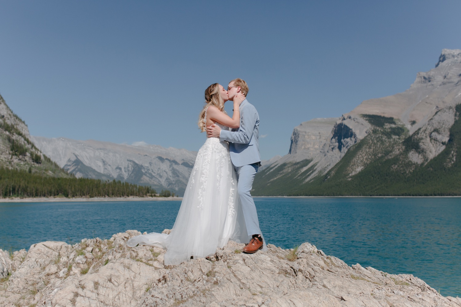 Groom in dusty blue suit and bride wearing fine detailed wedding dress kissing during midday portraits at Lake Minnewanka